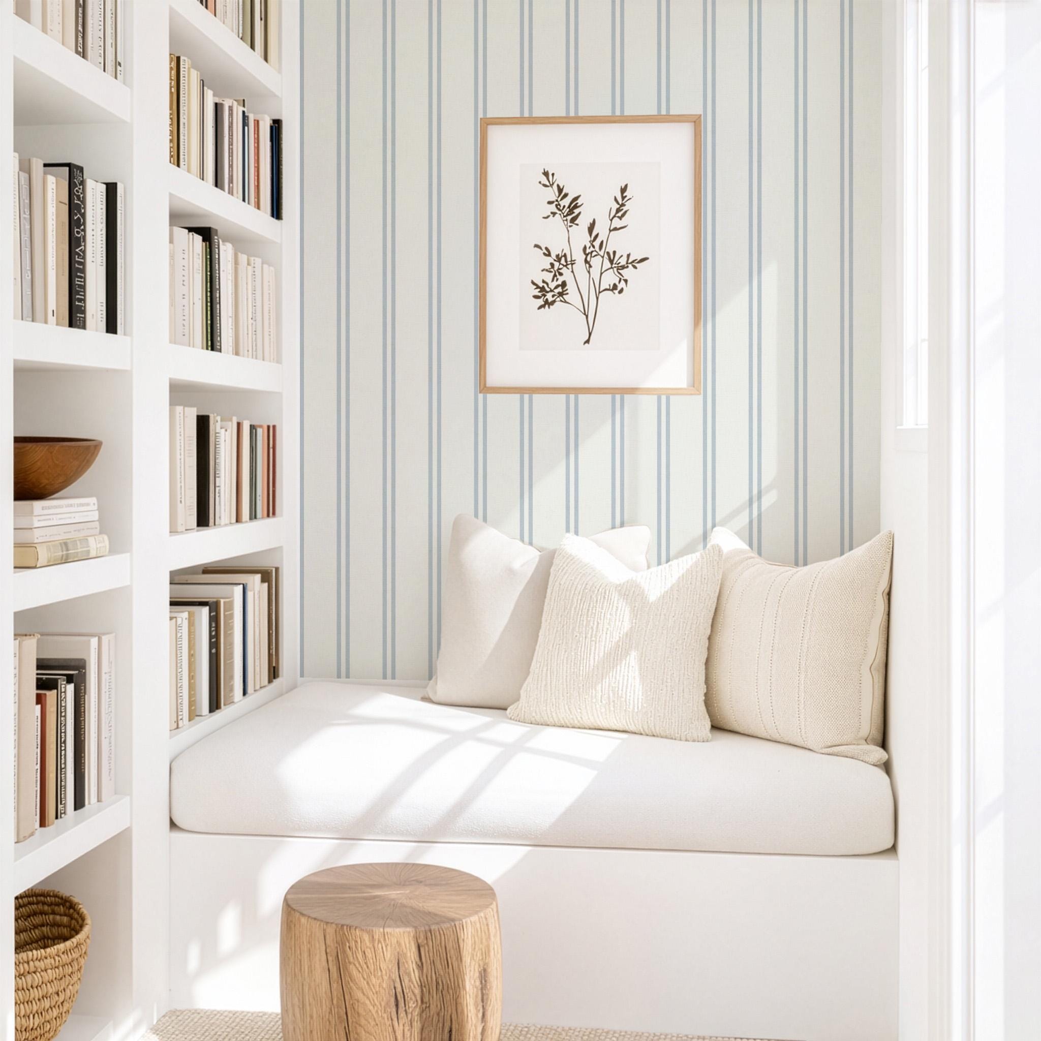May include: A cozy reading nook with a white built-in bench, three cream-colored pillows, and a wooden side table. A framed botanical print hangs on the striped blue and white wallpaper. Bookshelves are on the left.