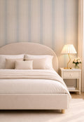 May include: A beige upholstered bed with a curved headboard, adorned with pillows and a blanket. A nightstand with a lamp and flowers sits beside the bed. The room has striped wallpaper and a neutral color palette, creating a serene bedroom setting.