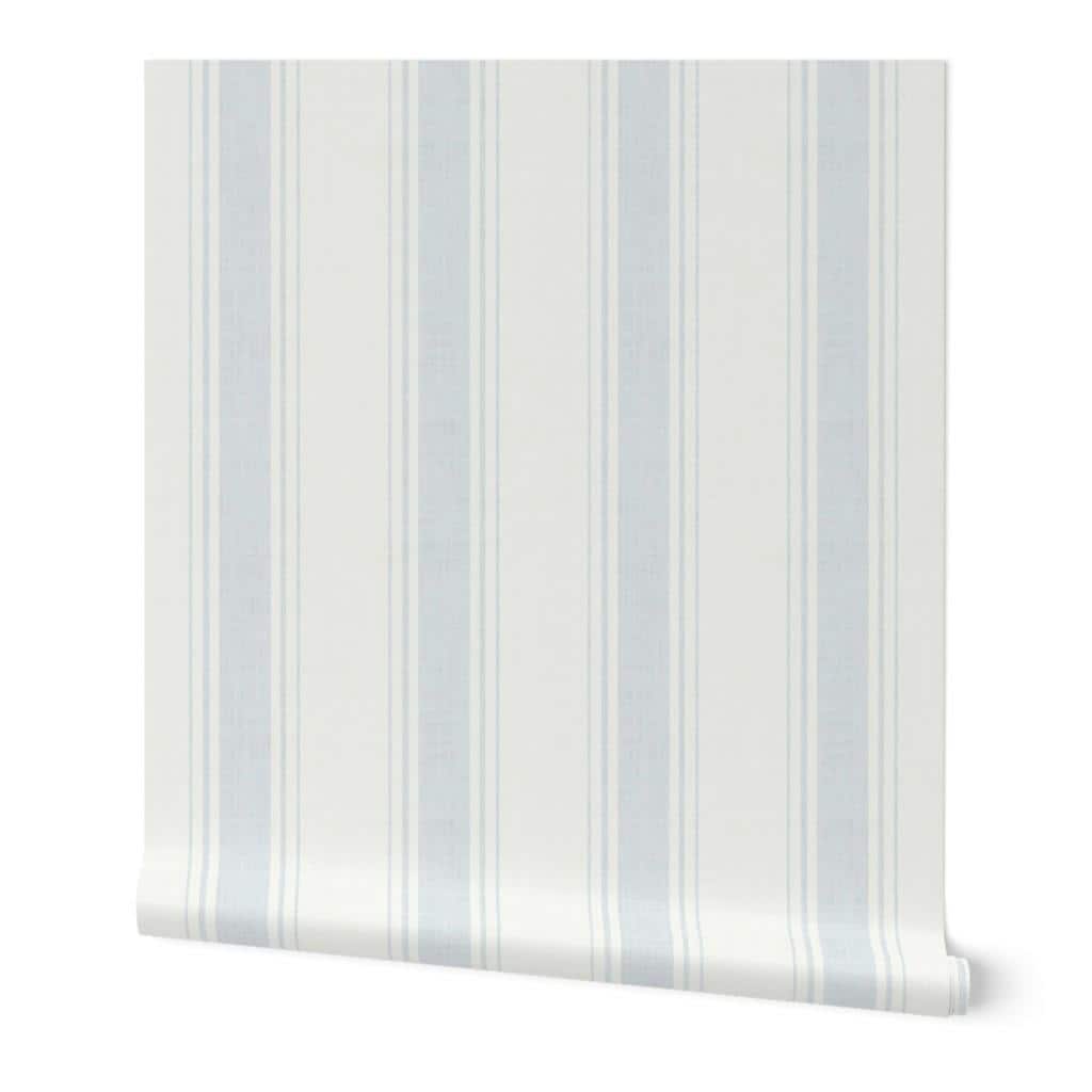 May include: A beige upholstered bed with a curved headboard, adorned with white and beige pillows and a matching throw. A bedside table with a gold lamp and a vase of flowers sits beside the bed. The wall has a striped pattern.