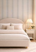 May include: A beige upholstered bed with a curved headboard, adorned with pillows and a blanket. A bedside table with a lamp and flowers sits beside the bed. The wall features vertical stripes in beige and light blue.