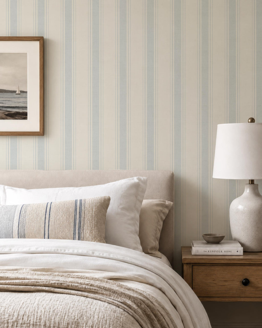 May include: A bedroom scene featuring a bed with white linens, pillows, and a beige throw. A striped pillow with blue stripes adds a decorative touch. A wooden nightstand holds a lamp and a book. The wall has a striped pattern.