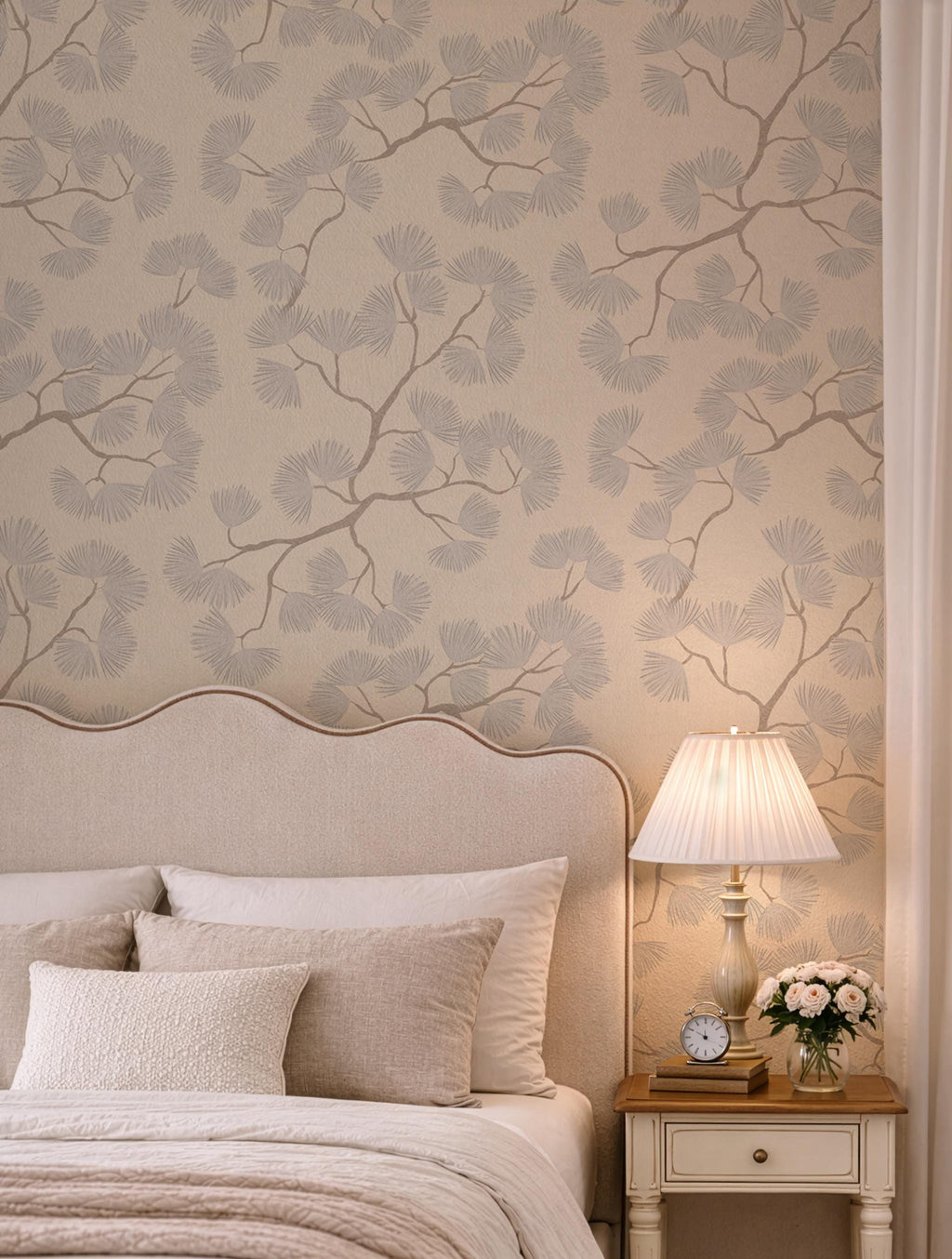 May include: A bedroom scene featuring a beige upholstered headboard, pillows, and bedding. A nightstand holds a lamp, clock, and flowers. The wallpaper has a repeating pattern of gray branches and leaves.