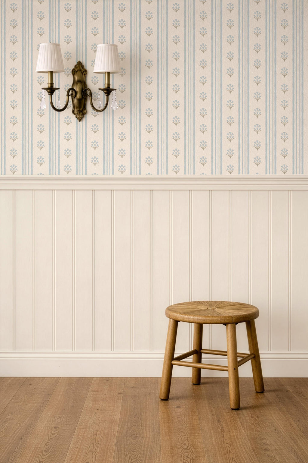 May include: A round wooden stool with a woven seat and four legs connected by a support frame, positioned against a wall with cream-colored wainscoting and wallpaper featuring vertical blue stripes and small floral patterns, with a wall sconce.